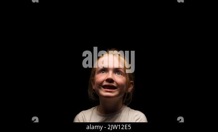 Junge kaukasische Frau mit einem glücklichen Ausdruck, der in einer dunklen Umgebung nach oben blickt. Stockfoto