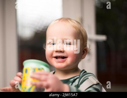 Junges Kleinkind hält während des Abendessens einen Becher und blickt mit einem netten Lächeln in die Kamera. Stockfoto