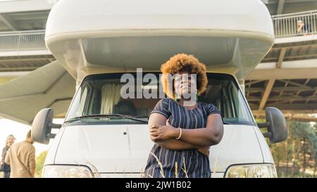 Dunkelhäutig lächelnde afrikanische junge Frau mit verkochten Haaren vor ihrem bequemen weißen Wohnmobil. Hochwertige Fotos Stockfoto