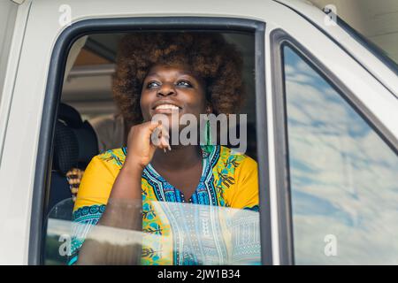 Dunkelhäutig lächelnde afrikanische junge Frau mit Afro-Haarschnitt, in einem hellen Boho-Outfit, die aus dem Fenster eines Wohnwagens blickt und die Aussicht während ihrer langen Reise um die Welt bewundert. Hochwertige Fotos Stockfoto