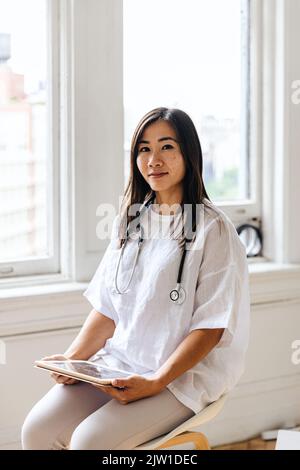 Porträt eines selbstbewussten jungen Arztes, der mit einem digitalen Tablet in der Klinik sitzt Stockfoto