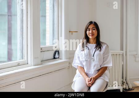 Porträt einer selbstbewussten Ärztin, die in der Klinik am Fenster sitzt Stockfoto