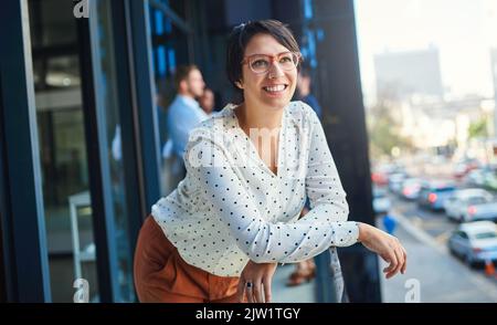 Die Zukunft sieht hell aus, von wo aus ich stehe. Eine junge Geschäftsfrau steht auf einem Balkon mit ihren Mitarbeitern im Hintergrund. Stockfoto