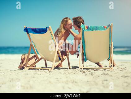 Komm her, du. Rückansicht eines jungen Paares, das auf Liegestühlen am Strand sitzt. Stockfoto