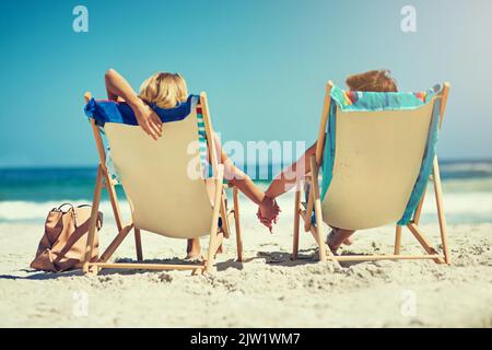 Seite an Seite am Meer. Rückansicht eines jungen Paares, das Hand in Hand auf Liegestühlen am Strand sitzt. Stockfoto