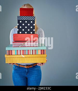 Hinter diesem Stapel von Geschenken. Beschnittene Studioaufnahme einer jungen Frau, die einen Stapel Geschenke vor ihrem Gesicht hält. Stockfoto