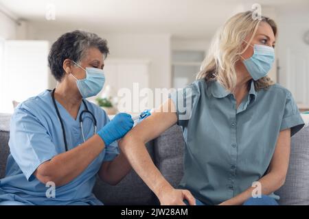 Ältere afroamerikanische Ärztin in Gesichtsmaske, die weibliche Patientin zu Hause impft Stockfoto