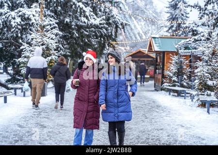 Zwei niedliche Mädchen laufen im Winter durch die Weihnachtsstadt. Frauen auf dem Hintergrund einer festlich verschneiten Stadt. Freunde Stockfoto