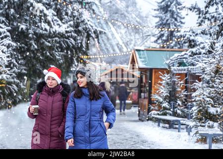 Zwei niedliche Mädchen laufen im Winter durch die Weihnachtsstadt. Frauen auf dem Hintergrund einer festlich verschneiten Stadt. Freunde Stockfoto