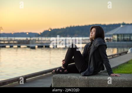 Lächelndes Teenager-Mädchen oder junge Erwachsene Frau in grauer Jacke sitzen im Freien am See und genießen den farbenfrohen Sonnenuntergang an einem kühlen Abend Stockfoto