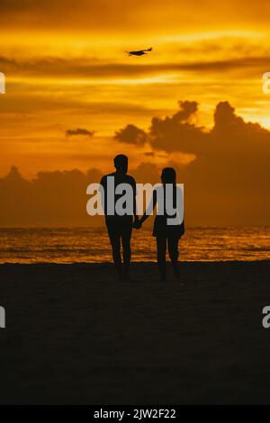 Rückansicht Silhouette eines anonymen liebenden Paares, das die Hände hält und am Strand in Richtung Wellenmeer geht, gegen den malerischen Sonnenuntergang in Clearwater Stockfoto