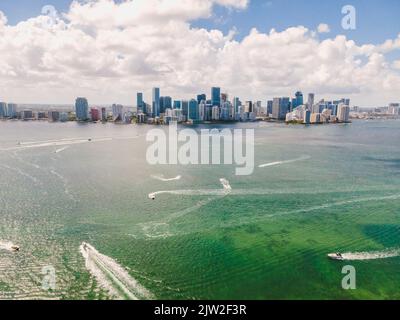 Atemberaubende Drohnenansicht verschiedener Motorboote, die in der Nähe moderner Wolkenkratzer im Meer schweben, gegen den wolkigen blauen Himmel in Miami Beach Stockfoto