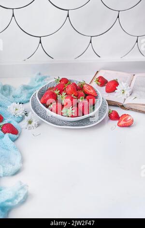 Von oben Schüssel mit reifen Erdbeeren auf der Theke in der Nähe von frischen weißen Blumen platziert und öffnete Buch tagsüber zu Hause Stockfoto