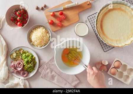 Draufsicht auf eine unkenntliche Frau, die beim Zubereiten von Quiche in der Küche im Ho mit einem Schneebesen rohe Hühnereier in einer Schüssel in der Nähe von frischen Zutaten und Kruste schlug Stockfoto