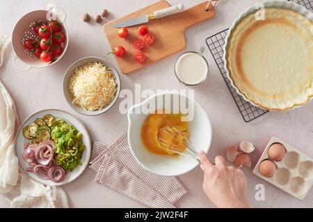 Draufsicht auf eine unkenntliche Frau, die beim Zubereiten von Quiche in der Küche im Ho mit einem Schneebesen rohe Hühnereier in einer Schüssel in der Nähe von frischen Zutaten und Kruste schlug Stockfoto