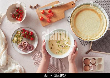 Von oben anonyme Frau, die Schüssel mit rohen Eiern mit Milch und Gemüse in der Nähe von leerer Kruste trägt, während sie Quiche zum Mittagessen in der Küche zu Hause kocht Stockfoto