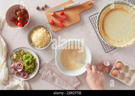 Draufsicht auf eine nicht erkennbare Frau, die Eier mit einem Schneebesen in der Schüssel in der Nähe von frischen Zutaten und Kruste schlug, während sie Quiche in der Küche zu Hause zubereitete Stockfoto