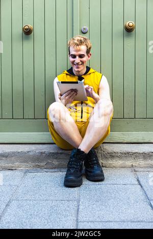 Ein junger lächelnder Transgender-Mann in gelbem Overall, der am Abend in der Nähe des Hauses auf der Straße der Stadt saß und auf dem Tablet browst Stockfoto