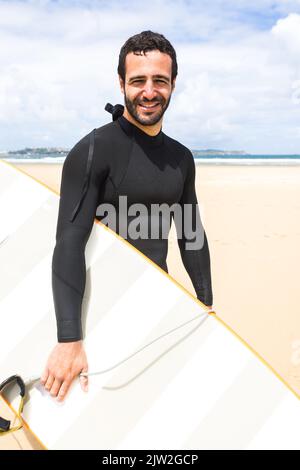 Fröhlicher junger hispanischer Surfer mit dunklem Haar und Bart in schwarzem Neoprenanzug, der lächelt, während er mit gelbem Surfbrett am Sandstrand steht Stockfoto