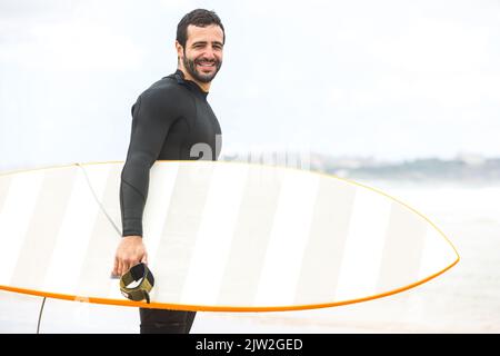 Seitenansicht eines fröhlichen jungen hispanischen männlichen Surfers mit dunklem Haar und Bart in schwarzem Neoprenanzug, der lächelt, während er mit weißem Surfbrett am Sandstrand steht Stockfoto