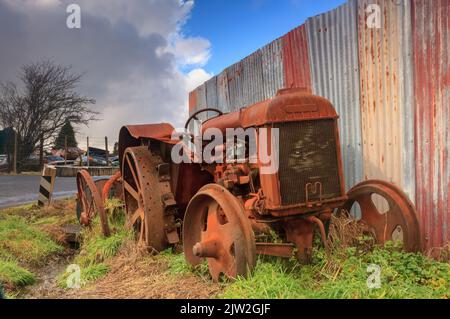 Ein alter, verrostet-out Traktor neben einem Wellblechzaun auf einem Wrackhof. Fotografiert im „Smash Palace“, Horopito, Neuseeland Stockfoto