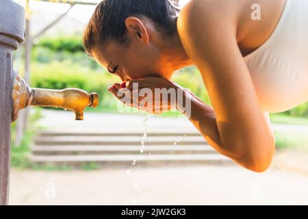 Seitenansicht von fit weiblich in activewear Trinkwasser aus öffentlichen Wasserhahn während Outdoor-Training im Park am Sommertag Stockfoto