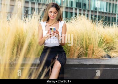 Seriöse junge Frau mit langen braunen Haaren in stilvollem Oberteil und Rock auf Steinbank in der Nähe von trockenem Gras sitzen und Messaging auf dem Smartphone im Stadtpark Stockfoto