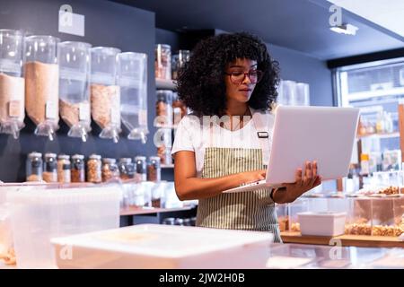 Junge afroamerikanische Frau in Uniform und Brille mit lockigem Haar, die Waren in Glasschachteln überprüft und einen Laptop benutzt, während sie in einem Zero Waste Lebensmittelgeschäft arbeitet Stockfoto
