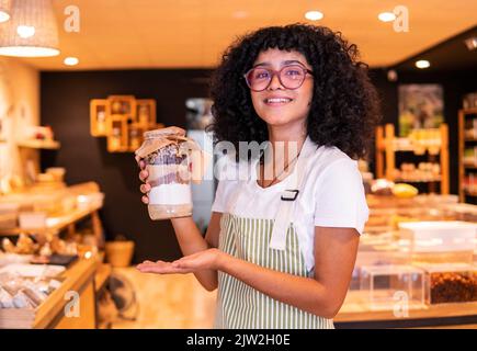 Glückliche junge ethnische Frau in Uniform und Brille lächelnd hält Glasglas mit Nüssen und verschiedenen Mehlen zur Kamera, während sie in Zero Waste Lebensmittelgeschäften arbeitet Stockfoto