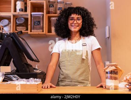 Junge lächelnde afroamerikanische Verkäuferin mit Schürze und Brille, die während der Arbeit in Zero Waste gr. auf die Kamera in der Nähe der modernen Registrierkasse schaut Stockfoto