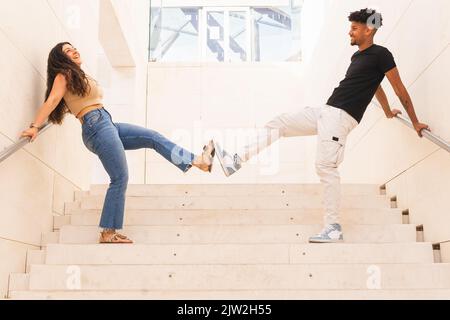 Das ganze lächelnde junge Paar berührt die Zehen, während es auf einer Treppe im Gebäude steht Stockfoto