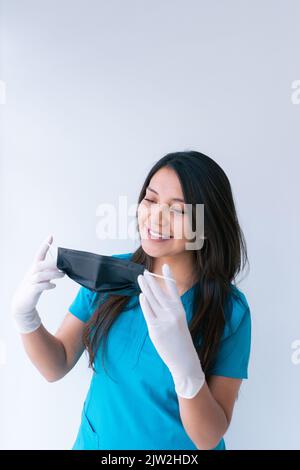 Fröhliche junge ethnische Ärztin mit langen dunklen Haaren in blauer medizinischer Uniform und Latexhandschuhen, die eine Schutzmaske aufsetzen und gegen Weiß lächeln Stockfoto