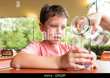 Unverkennbarer Elternteil, der sauberes Wasser aus dem Krug in das Glas des staunenden Sohnes goss, während er Zeit im Restaurant im Park verbrachte Stockfoto