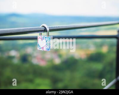 Das verriegelte Schloss hängt am Metallzaun. Symbol der Liebe im Park. Das metallrote Schloss in Herzform ist mit einem Schlüssel verschlossen Stockfoto