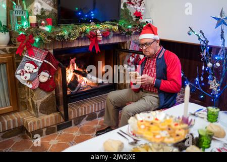 Fokussierter reifer Mann in Santa hat SMS-Nachricht auf Handy, während in der Nähe dekorierten Kamin während der Weihnachtsfeier im Wohnzimmer sitzen Stockfoto