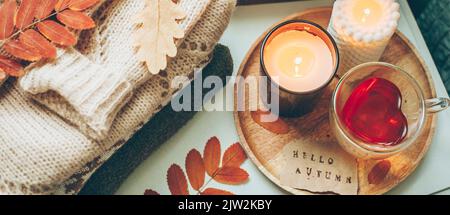 Stapel gestrickter Pullover, Kerzen, Tasse mit Tee und Herbstblättern. Konzept des gemütlichen Wohnraums, Stockfoto