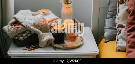 Konzept der Stillleben im Herbst. Gemütliche Komposition mit brennenden Kerzen, einer Tasse Tee aus Glas, warmen Pullovern auf dem Betttisch im Schlafzimmer. Stockfoto
