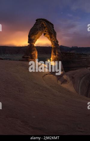 Nicht erkennbarer Reisender, der mit Taschenlampe in der Nähe der Natursteinformation Delicate Arch in der trockenen Wüste gegen den wolkigen Sonnenuntergang I steht Stockfoto