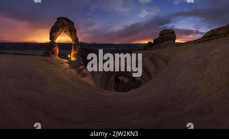 Nicht erkennbarer Reisender, der mit Taschenlampe in der Nähe der Natursteinformation Delicate Arch in der trockenen Wüste gegen den wolkigen Sonnenuntergang I steht Stockfoto