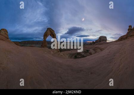 Atemberaubende Aussicht auf Delicate Arch in der Nähe einer rauen Schlucht gegen bewölkten Himmel mit Mond im Arches National Park in Utah, USA Stockfoto