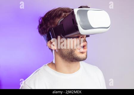 Staunend männlich im lässigen T-Shirt modernes VR-Headset erleben Cyberspace mit geöffnetem Mund, während auf buntem Hintergrund im Studio stehen Stockfoto