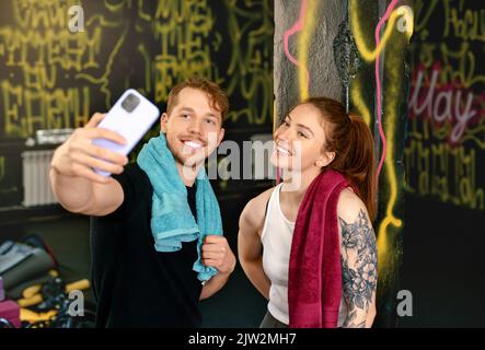 Fröhlicher junger Sportler und Sportlerin im aktiven Ohr lächelnd und Selfie auf dem Handy während der Pause im Fitnesstraining im Fitnessstudio Stockfoto