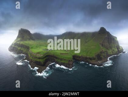 Atemberaubende Drohne Blick auf die Insel mit rauen felsigen Klippen mit grünem Gras bedeckt mit Siedlung Dorf umgeben von winkenden Meer gegen graue Klo Stockfoto