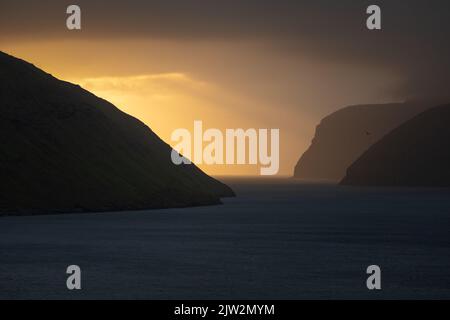 Atemberaubende Aussicht auf das Meerwasser, das in der Nähe von Klippen vor dem wolkigen Sonnenaufgangshimmel am frühen Morgen auf den Färöer Inseln plätschern kann Stockfoto