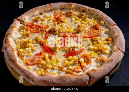 Pizza für Kinder auf Sahnebasis mit Hühnerfilet, Tomaten, Mais und Mozzarella-Käse auf schwarzem Hintergrund. Stockfoto