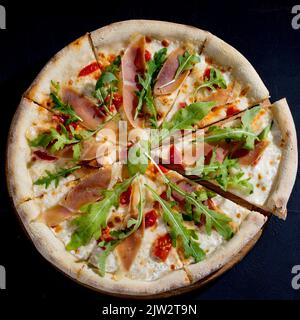 Pizza mit Jamon oder Prosciutto, Mozzarella, Kirschtomaten, Rucola und Parmesan auf schwarzem Hintergrund. Italienische Küche Stockfoto