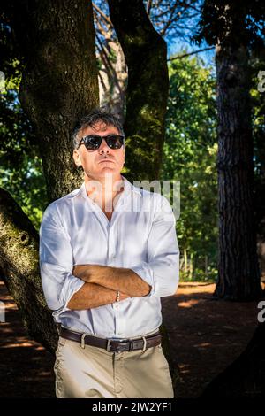 Der junge Mann trägt eine Sonnenbrille, ein weißes Hemd und eine Bedge-Hose. Ein gutaussehender Geschäftsmann arbeitet im Park. Stockfoto