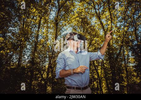 Der junge Mann nutzt einen Virtual-Reality-Betrachter im Park. Der gutaussehende Geschäftsmann arbeitet mit einem Virtual-Reality-Betrachter im Freien. Stockfoto