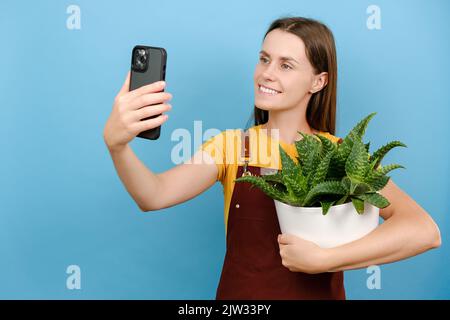 Lächelnd nette Frau Floristen blogger mit Pflanzen im Topf auf Smartphone-Kamera fotografiert, isoliert über blauen Hintergrund Wand. Gartenarbeit, Housewif Stockfoto