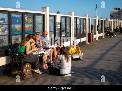 Eine Familiengruppe sitzt am Strand am Worthing Pier, West Sussex, Großbritannien, um traditionellen Fisch und Chips zu essen. Stockfoto